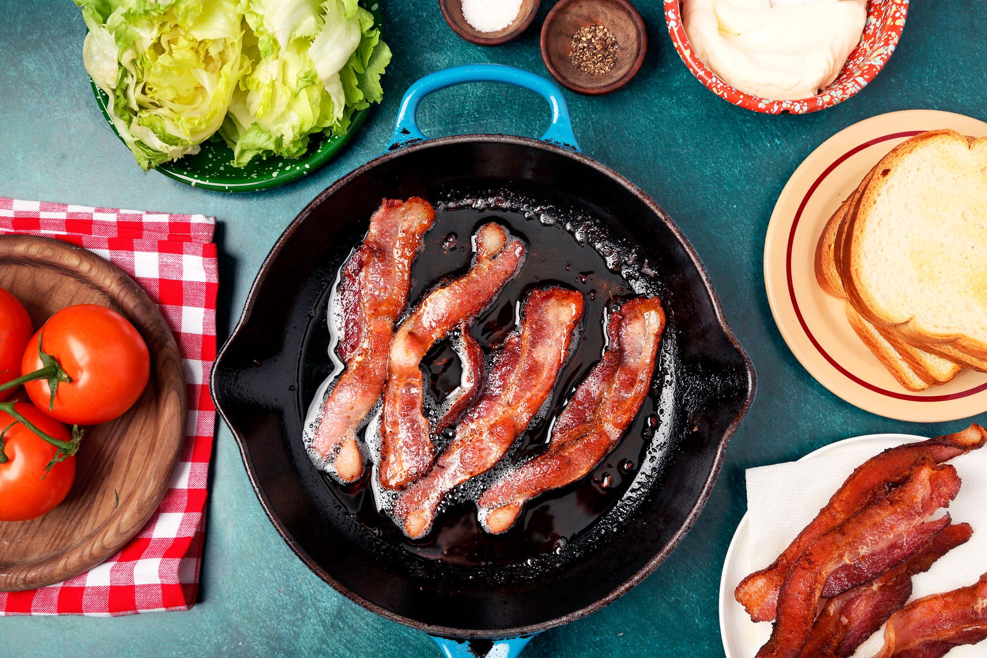 overhead shot of a cast iron skillet filled with crispy bacon is at the center of the composition, surrounded by various other ingredients