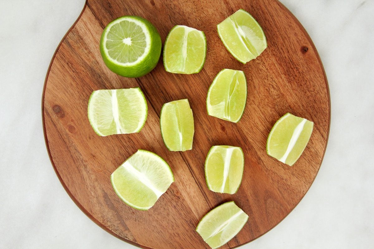 A wooden board with ten lime wedges and one half lime arranged on it, placed on a light countertop.