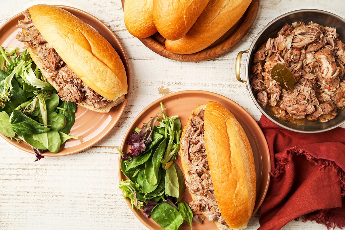 Sandwiches filled with shredded meat on plates with mixed greens. A bowl contains additional shredded meat, garnished with a bay leaf. More rolls are placed in the background on a wooden dish, all set on a light wooden table.