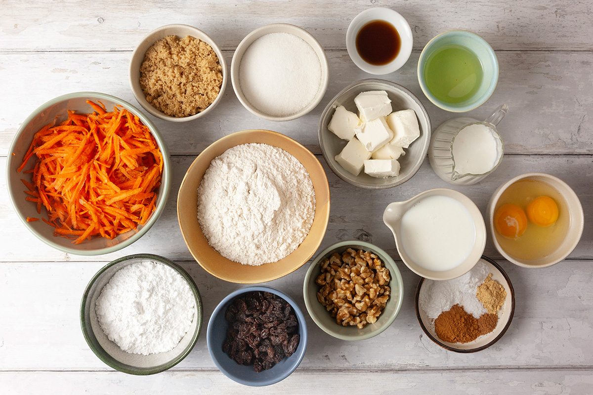 Top view of various baking ingredients in bowls on a white wooden surface, including shredded carrots, flour, sugar, eggs, butter, milk, oil, vanilla extract, nuts, spices, raisins, and baking powder.