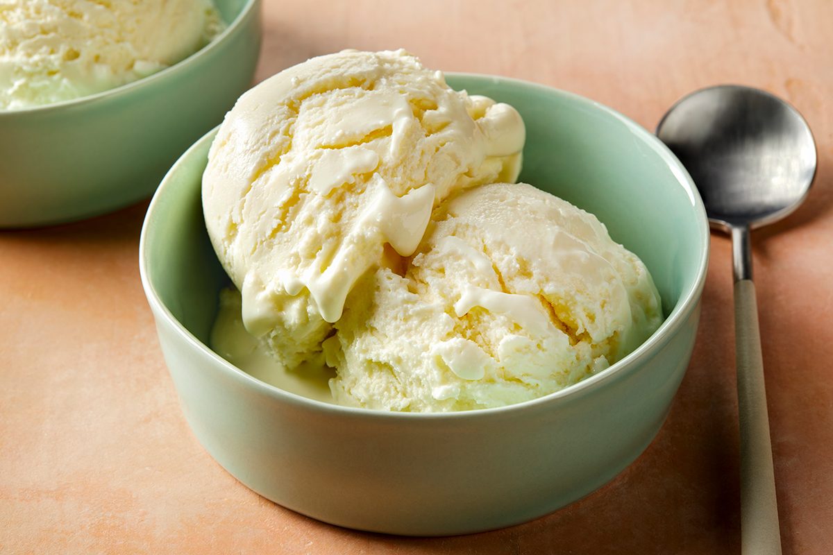 A teal bowl filled with two scoops of vanilla ice cream on a light brown surface. A spoon is placed next to the bowl. Another bowl of ice cream is partially visible in the background.