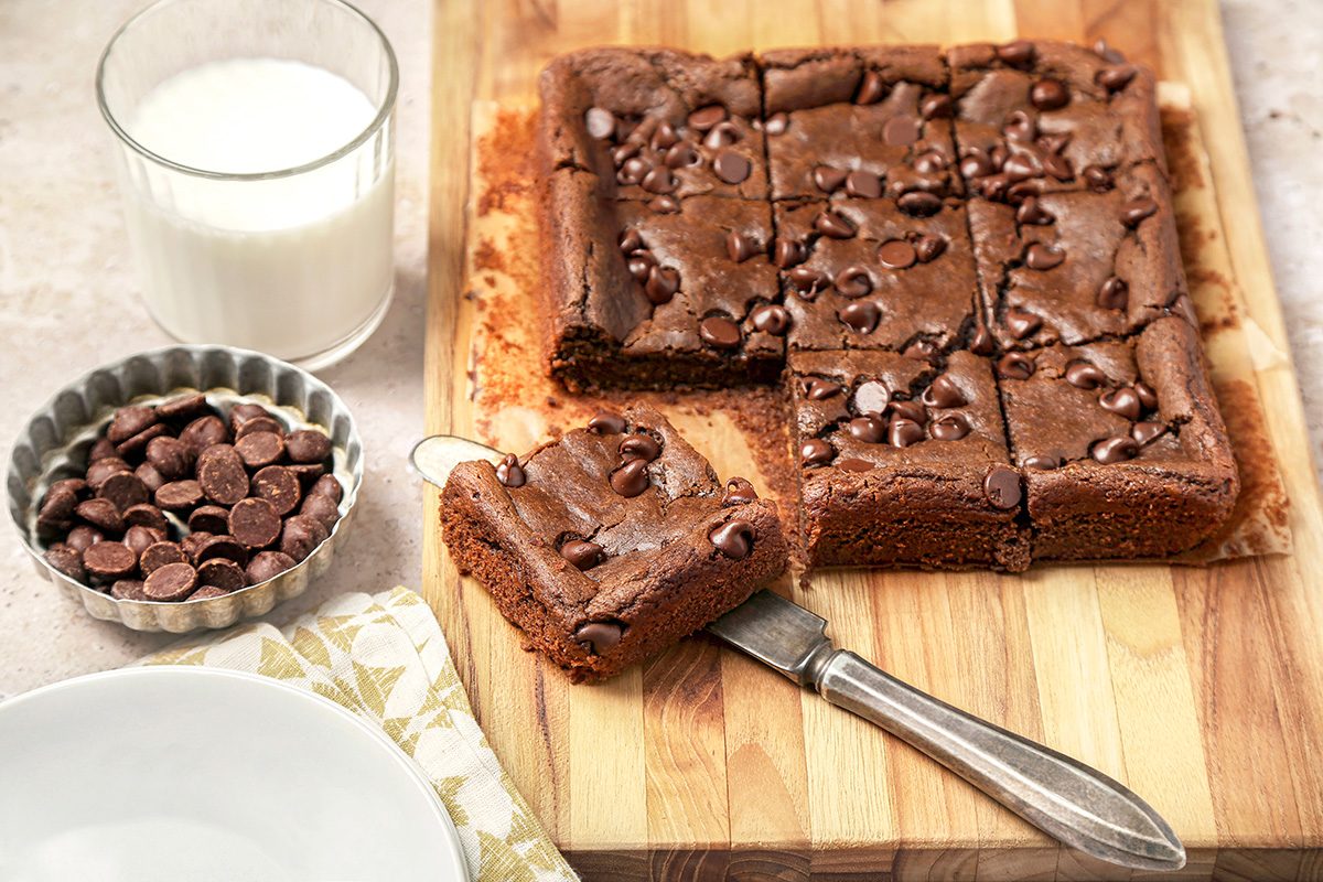 A batch of Chickpea chocolate brownies on a wooden board, cut into squares. One piece is on a knife. 