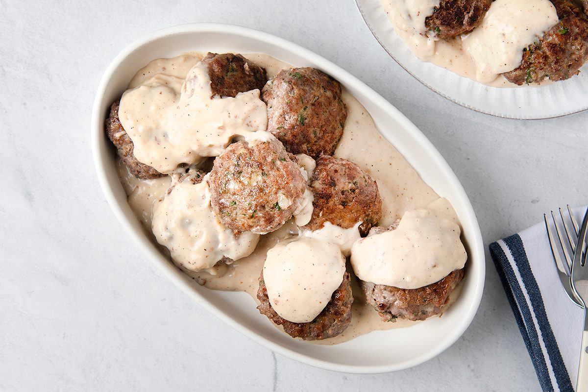 A white oval dish filled with cooked meat patties covered in creamy gravy. The dish is on a light gray surface, with part of a blue and white striped napkin visible at the corner.