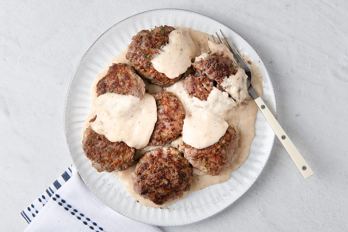 A white plate filled with seven beef patties topped with creamy white gravy. A fork rests on the plate's edge. The plate sits on a light gray surface beside a folded white cloth with blue stripes.