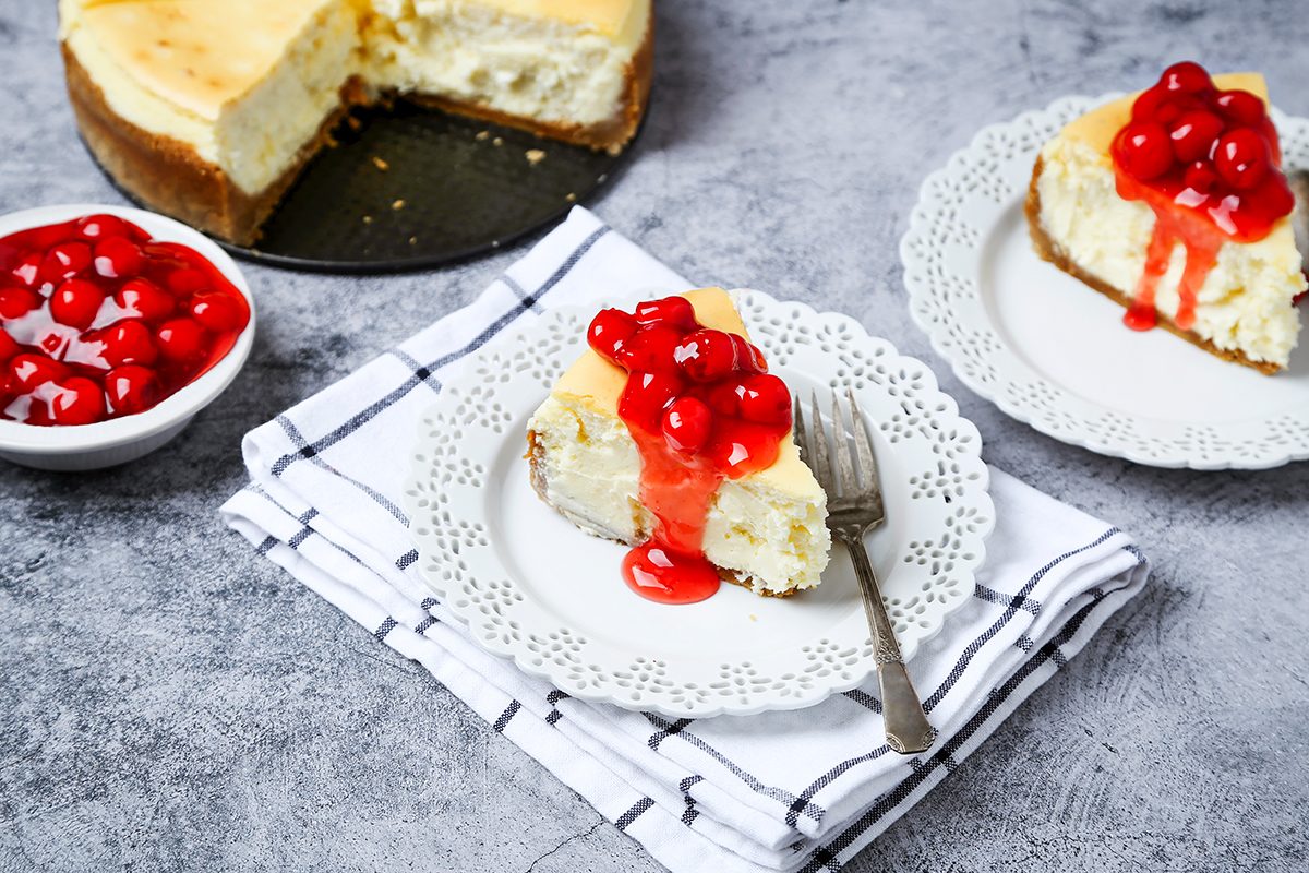 Two slices of cheesecake topped with cherries sit on decorative plates with forks. 
