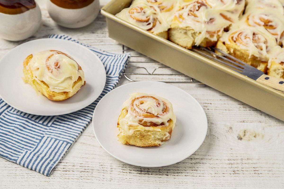 A white table displays a large baking dish filled with frosted cinnamon rolls. Two rolls are served on separate white plates, placed on a blue striped cloth. Jars are visible in the background.