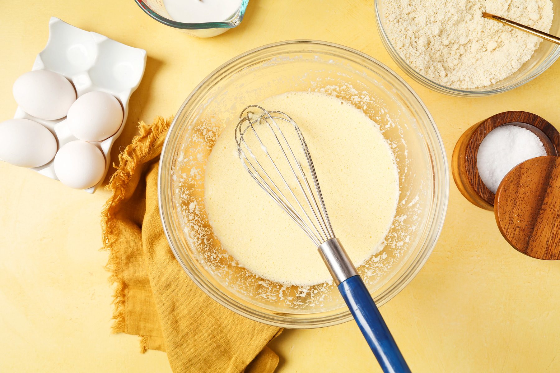 Keto Waffles shot on a yellow surface. Waffler batter mixed in a large mixing bowl with a whisk.