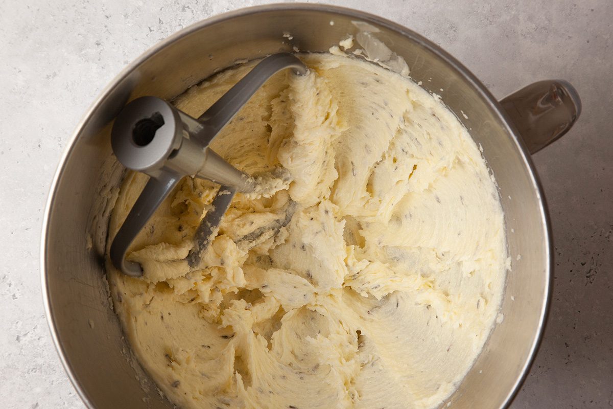 A metal mixing bowl filled with creamy, light beige dough or batter, with a paddle attachment. The mixture is smooth and evenly spread around the bowl. The bowl is set on a light-colored countertop.