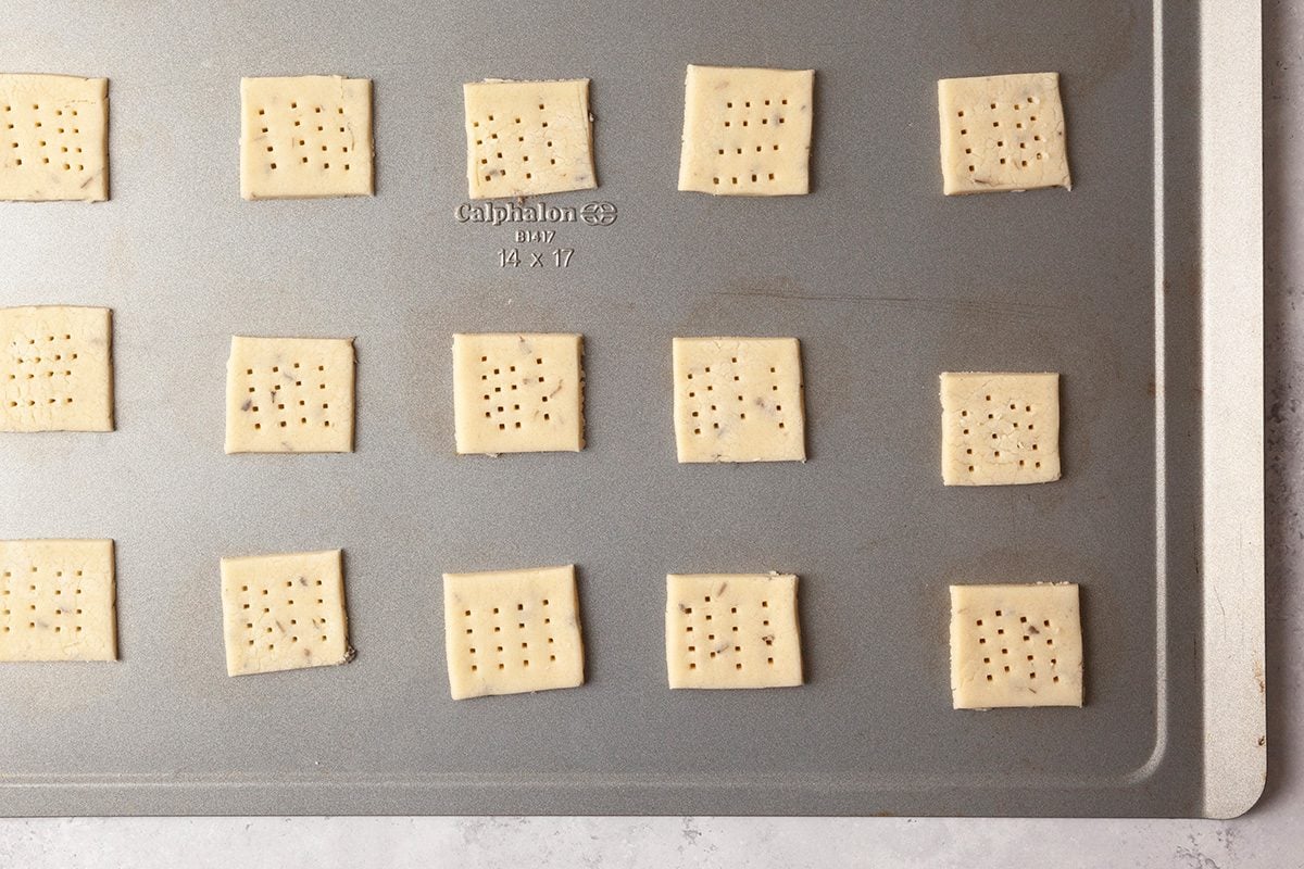 A baking tray with evenly spaced square cracker dough pieces, each pricked with small holes, ready for baking. The tray is on a light countertop.