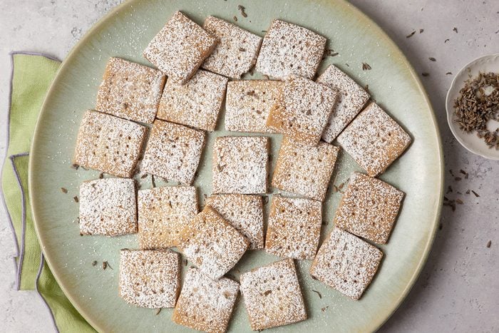 A large green plate holds square crackers dusted with a light powder. They are arranged neatly, with a small bowl containing seeds or spices beside the plate. A light green napkin is partially visible underneath.