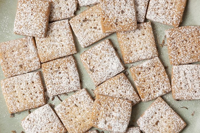 Square cookies dusted with powdered sugar are arranged in a scattered pattern on a light green surface. The cookies have a golden-brown color and a textured appearance.