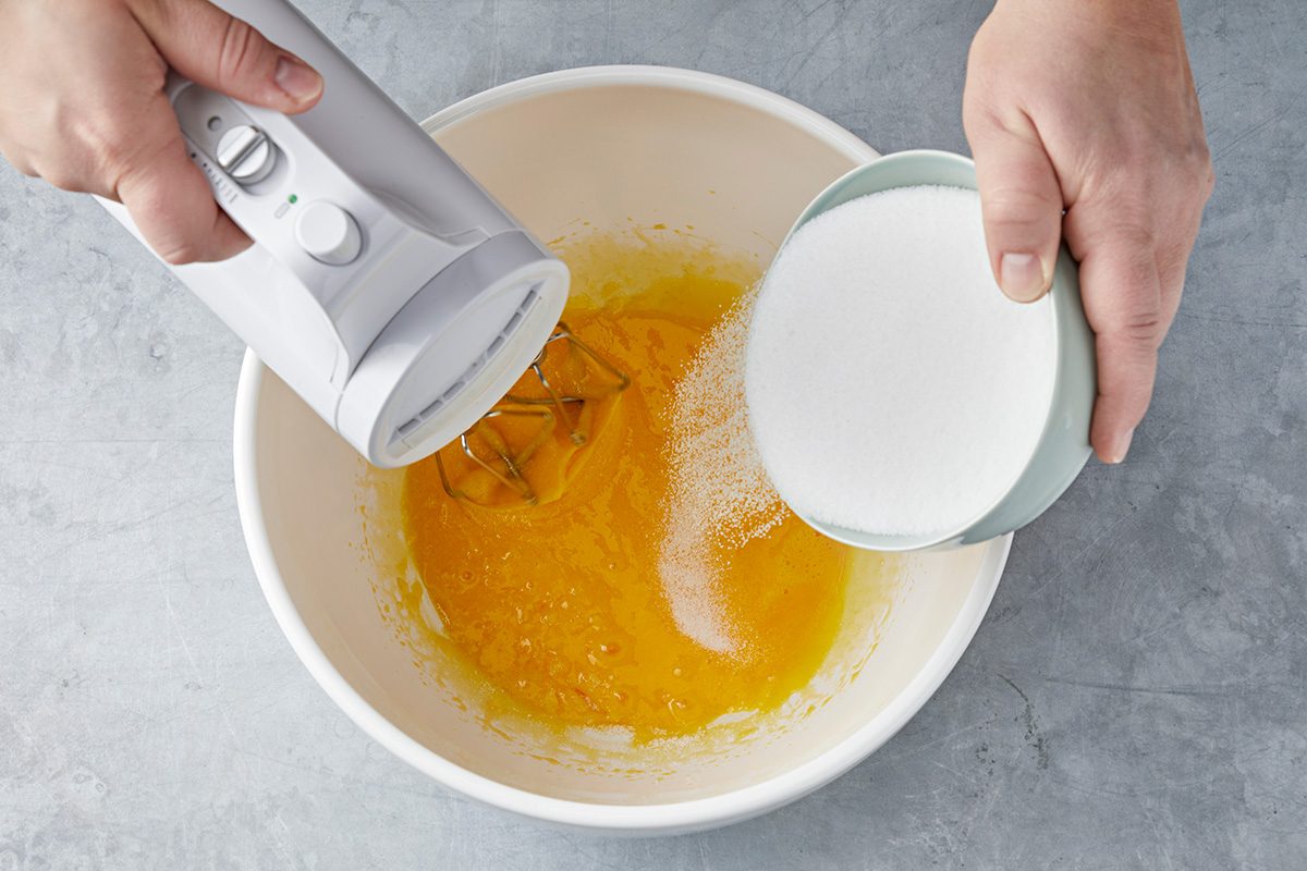 Hands hold a hand mixer and a bowl of sugar over a large mixing bowl containing a yellow liquid. The sugar is being poured into the bowl, likely for baking. The background is a light gray surface.