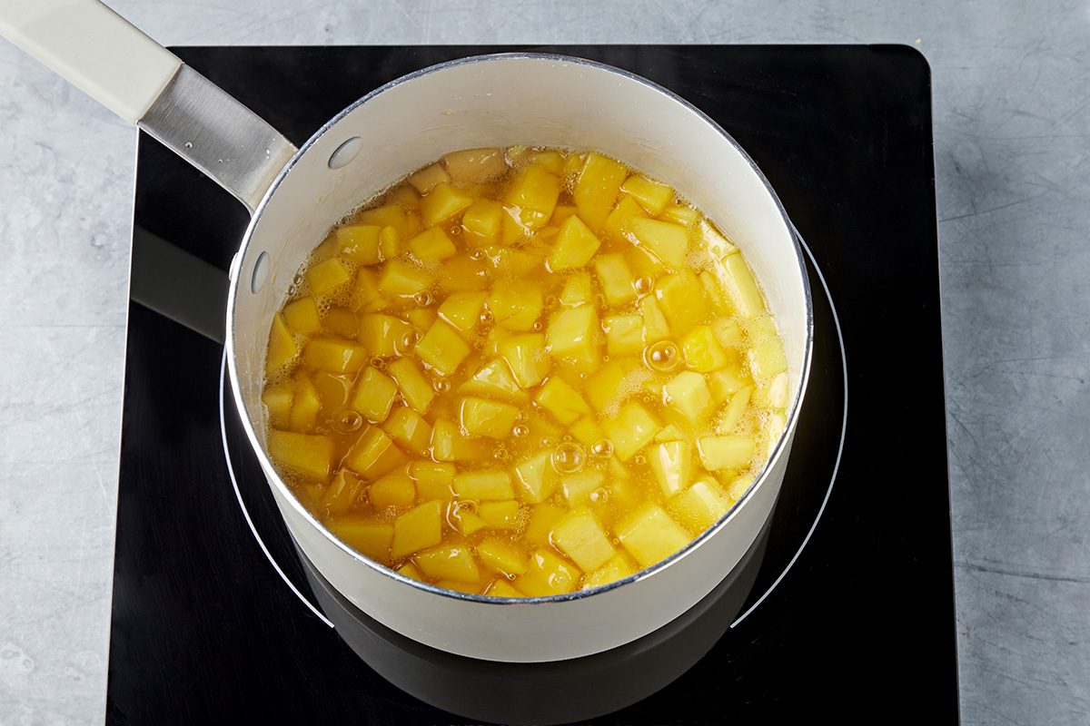 A pot filled with diced yellow mango pieces is cooking on a black stovetop. The mango chunks are simmering in liquid, appearing translucent and releasing steam. The stovetop is on a light-colored countertop.