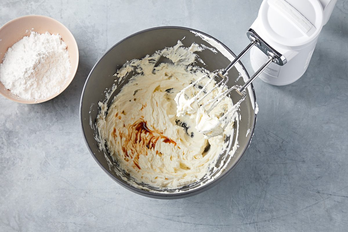 A mixing bowl with creamy batter and a splash of vanilla sits next to a hand mixer. A bowl of flour is placed nearby on a gray countertop.