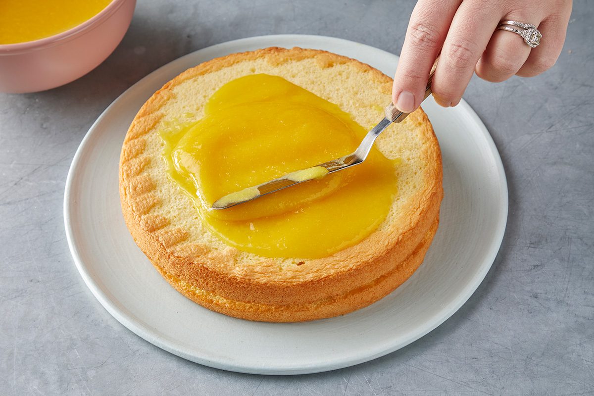 A hand spreads yellow lemon curd onto a round, layered sponge cake with a spatula on a white plate. A pink bowl can be seen in the background, placed on a gray countertop.