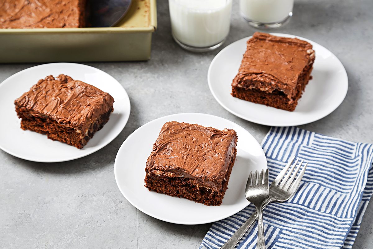 Three slices of chocolate frosted cake on white plates, placed on a gray surface. A striped blue and white cloth with two forks is nearby. In the background, a baking pan with more cake and two glasses of milk are visible.