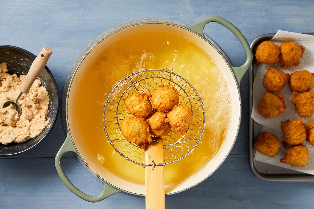 A pot of hot oil is used to fry golden hush puppies. A wire skimmer holds several cooked hush puppies above the pot. On the left is a bowl of uncooked batter, and on the right is a tray of cooked hush puppies on paper towels.