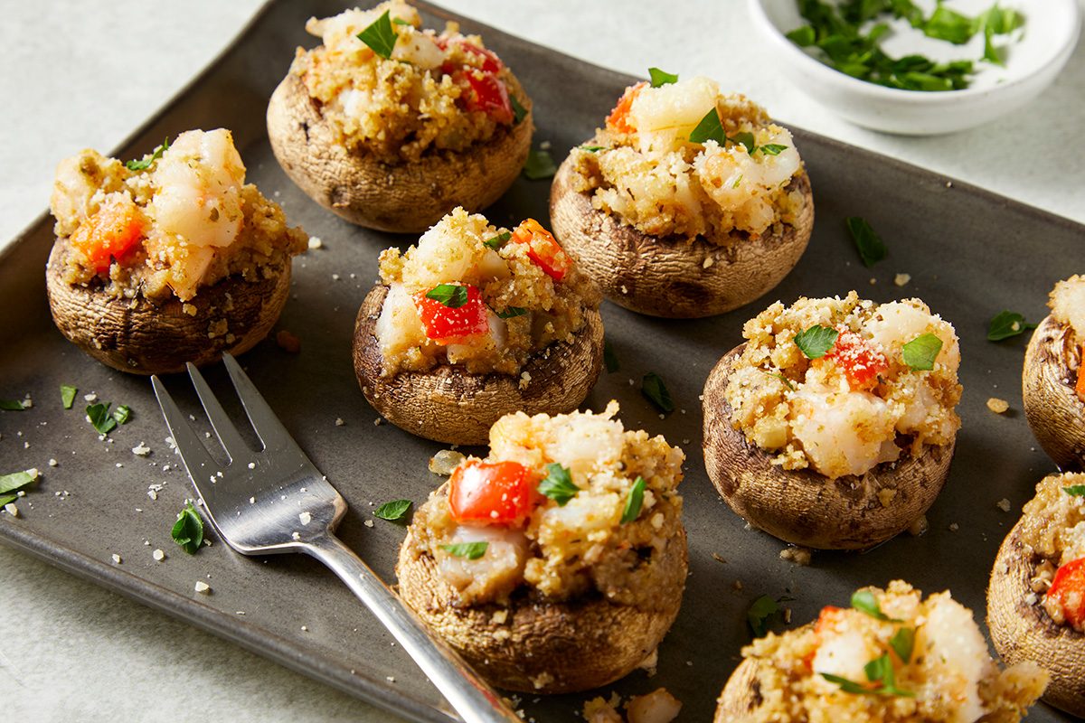 Stuffed mushrooms topped with breadcrumbs, diced vegetables, and herbs, arranged on a gray platter. A fork rests nearby. Finely chopped parsley is sprinkled on top, with a small bowl of additional parsley in the background.