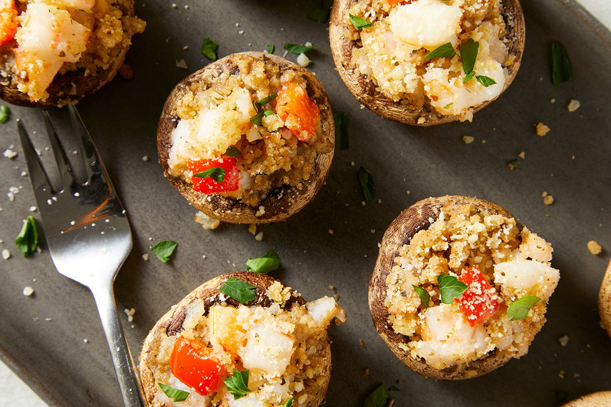 Stuffed mushrooms filled with a mixture of breadcrumbs, diced red peppers, and herbs, are arranged on a gray plate. A fork and scattered parsley leaves add to the presentation.