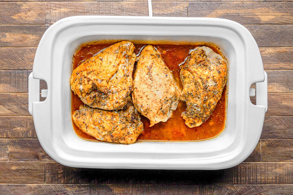 overhead shot of a white slow cooker on a wooden surface; the slow cooker is filled with four chicken breasts that have been cooked
