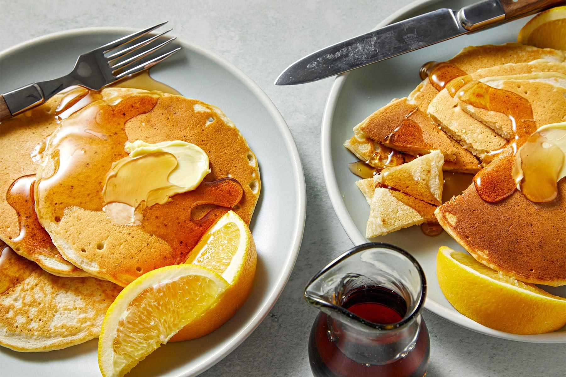 3/4th shot of sourdough pancakes served with a steel fork on two ceramic plates with butter, lemons and honey.