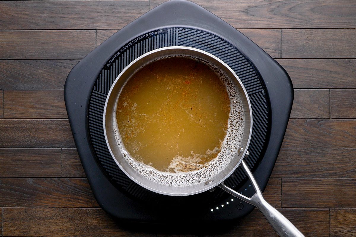 A pot of yellowish liquid, possibly broth or soup, sits atop an electric stovetop on a dark wooden surface. Bubbles are visible around the edges, and a metal ladle is placed inside the pot.