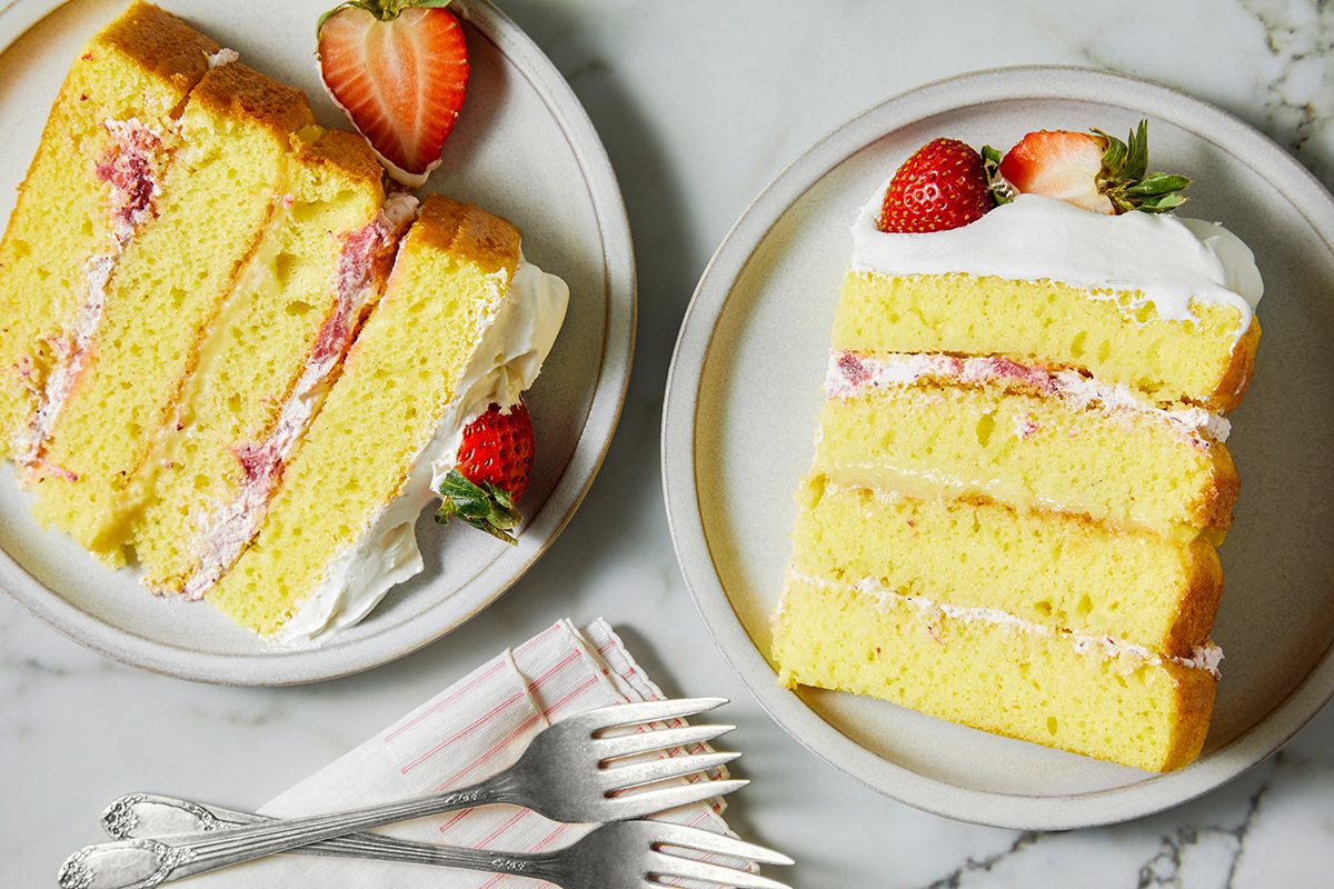 Two slices of layered yellow cake with white frosting and strawberry pieces on top sit on white plates. A whole strawberry garnishes each slice. Two metal forks rest on a napkin beside the plates, set on a marble surface.