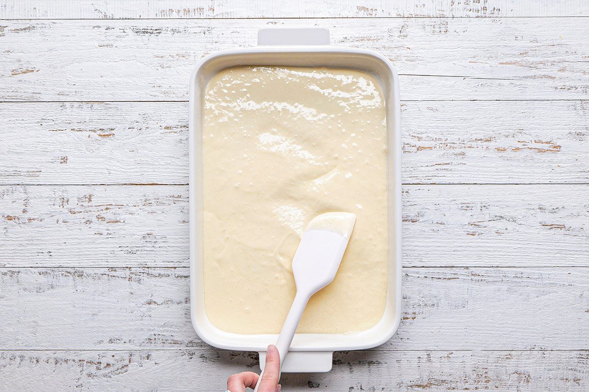 overhead shot of a rectangular baking dish filled with a smooth, creamy batter; a white spatula is used to spread the batter evenly within the dish; the dish is placed on a white wooden surface