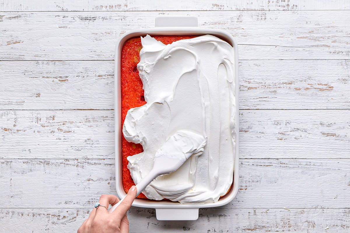 overhead shot of a rectangular baking dish filled with a red dessert topped with a thick layer of white whipped cream; a hand is seen using a spatula to spread the cream smoothly over the top; the background is a wooden countertop