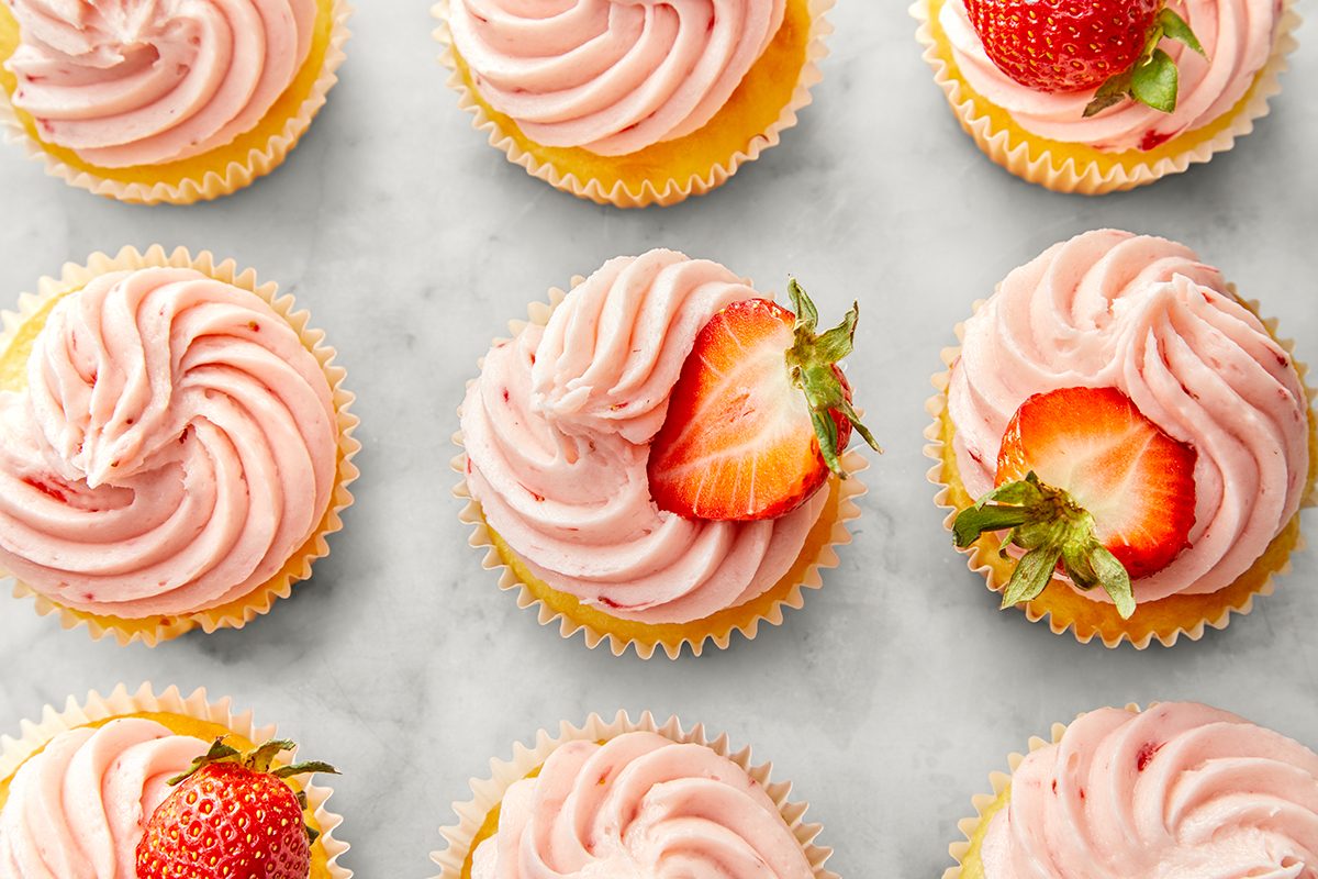 Cupcakes topped with swirls of pink frosting are arranged in rows on a light surface. Each cupcake is garnished with a halved strawberry, adding a touch of freshness to the tempting dessert display.