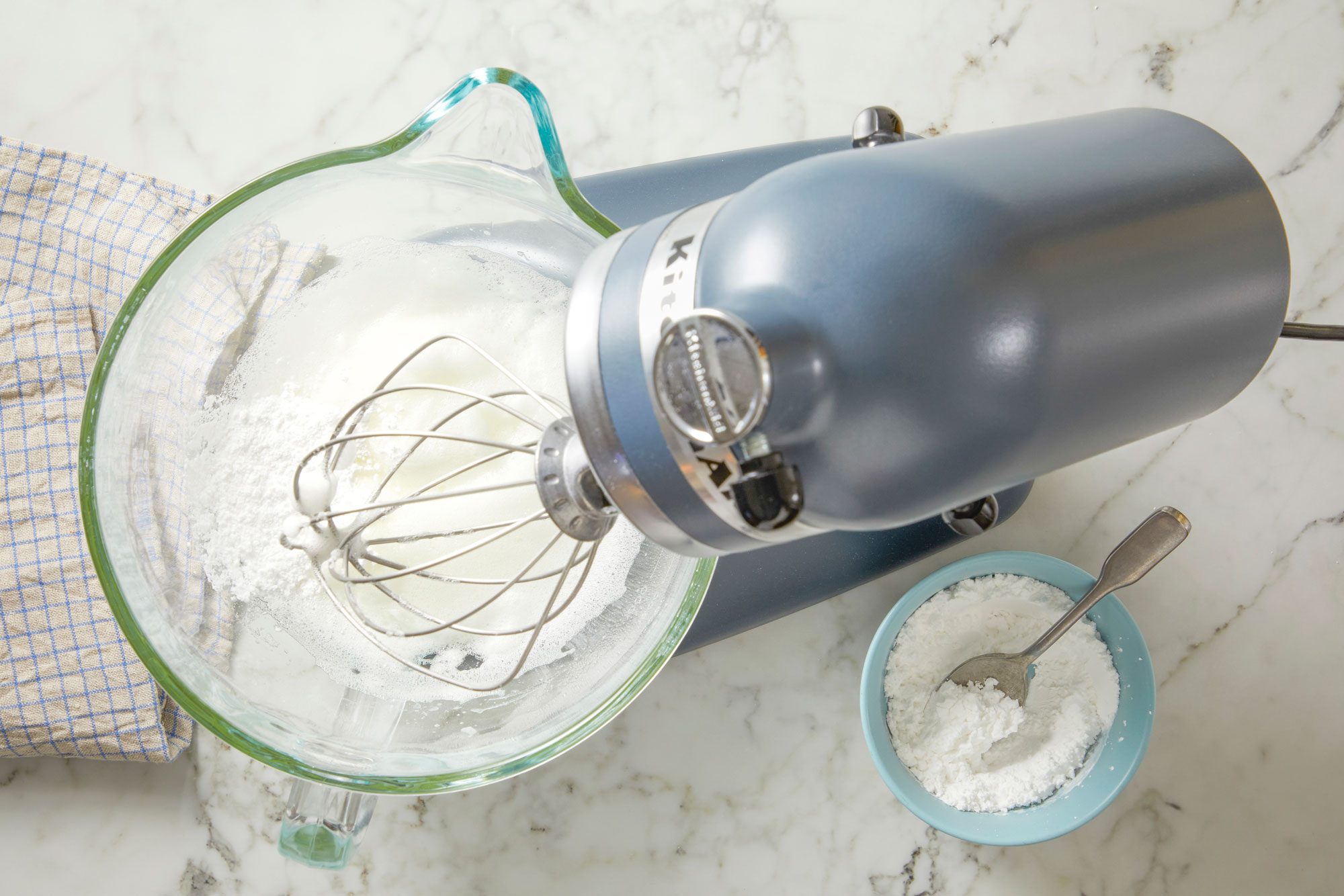 Overhead shot of place egg whites and salt in a very clean bowl of a stand mixer fitted with a whisk attachment; whisk on medium-low speed until frothy; Slowly add superfine sugar; whisk until dissolved 1-2 minutes; Slowly add remaining 1/2 cup confectioners’ sugar; increase speed to high and whip until meringue is glossy and stiff peaks form, 1-2 minutes; a napkin; on marble surface;