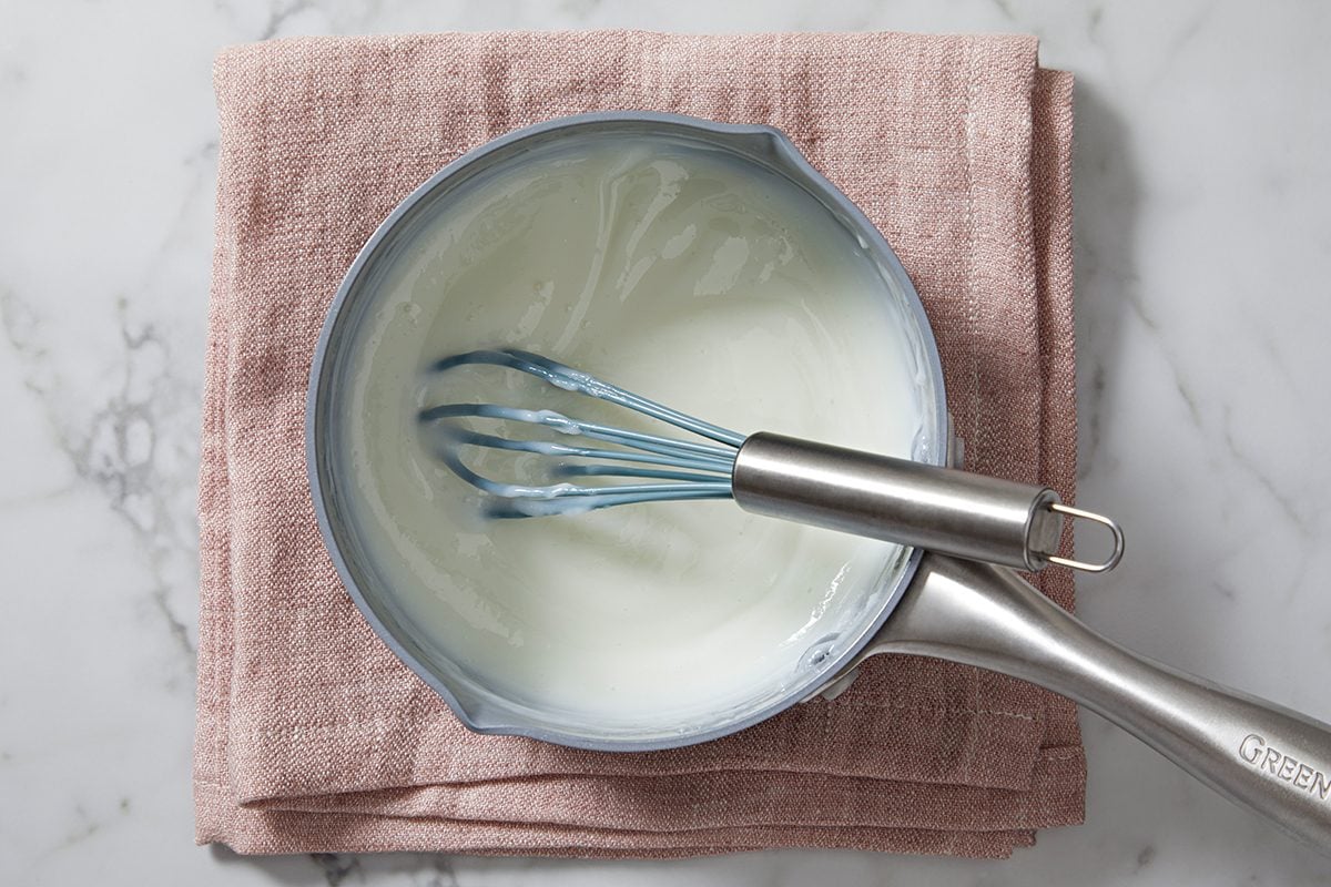A saucepan with creamy white sauce and a whisk inside sits on a folded pink fabric on a marble surface.