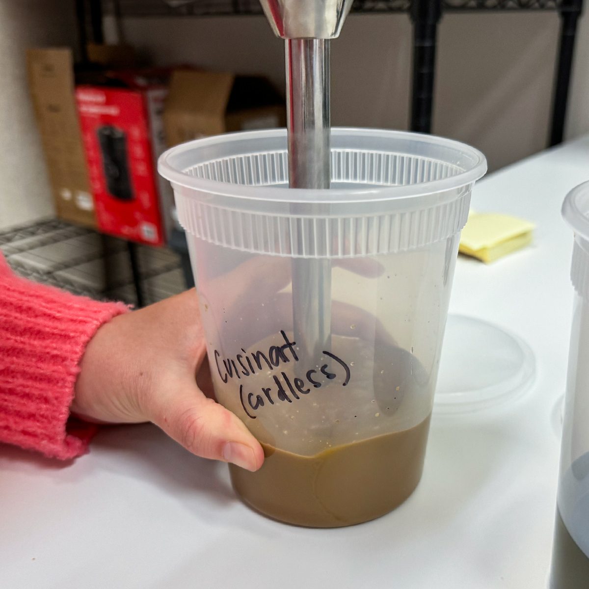 A person in a red sweater uses an immersion blender in a plastic container labeled "Cuisinart (cordless)," blending a brown liquid. The scene is set on a white countertop with shelves in the background.