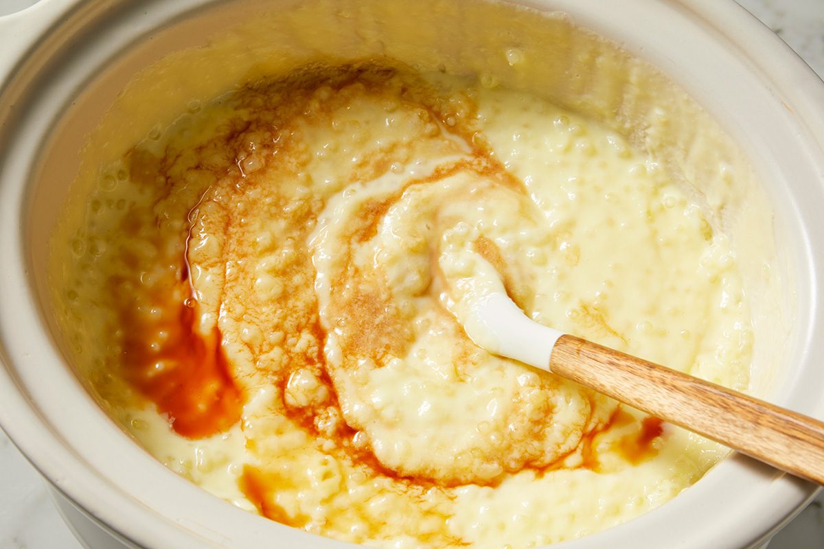 A slow cooker filled with creamy rice pudding, slightly browned on top. A wooden spoon rests inside, ready to stir the mixture. The pudding has a smooth texture with a golden hue.