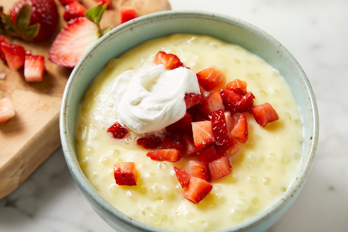 A bowl of creamy tapioca pudding topped with diced strawberries and a dollop of whipped cream, placed on a marble surface. Sliced strawberries are visible on a wooden board in the background.