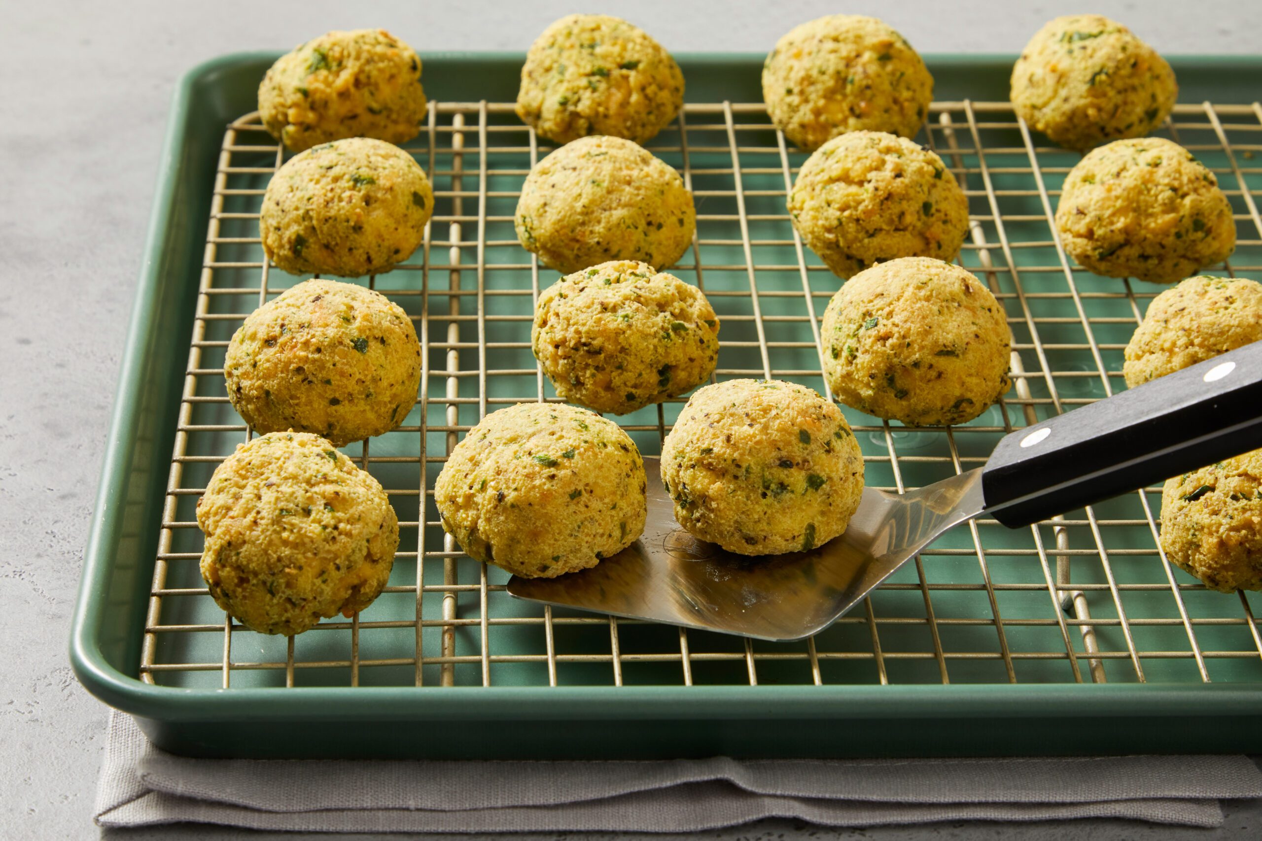 Golden-brown falafel balls are arranged on a cooling rack placed over a green baking sheet. A metal spatula on the right side holds one falafel. 