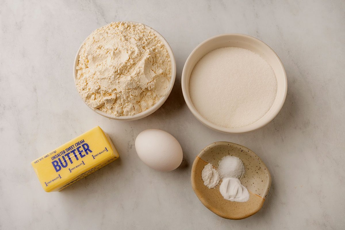 Birds-eye view of bowls of flour, sugar, and baking powders, a stick of butter, and an egg, on a white stone countertop