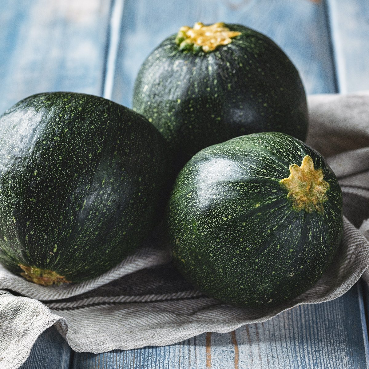 Three round, dark green zucchinis with light speckles and yellow stem tops rest on a gray cloth atop a rustic blue wooden surface.