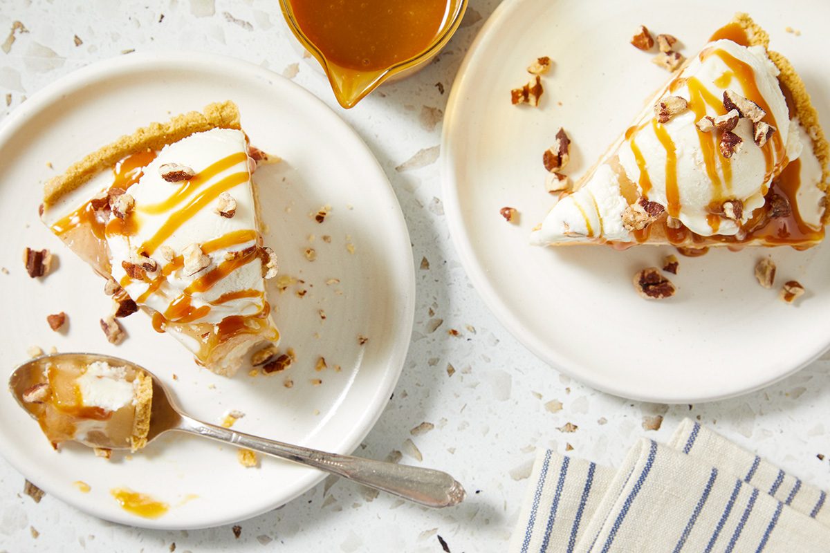 Two slices of pie topped with whipped cream, chopped pecans, and caramel sauce are served on white plates. A spoon rests by one slice, and a small pitcher of caramel sauce sits nearby on the speckled surface.