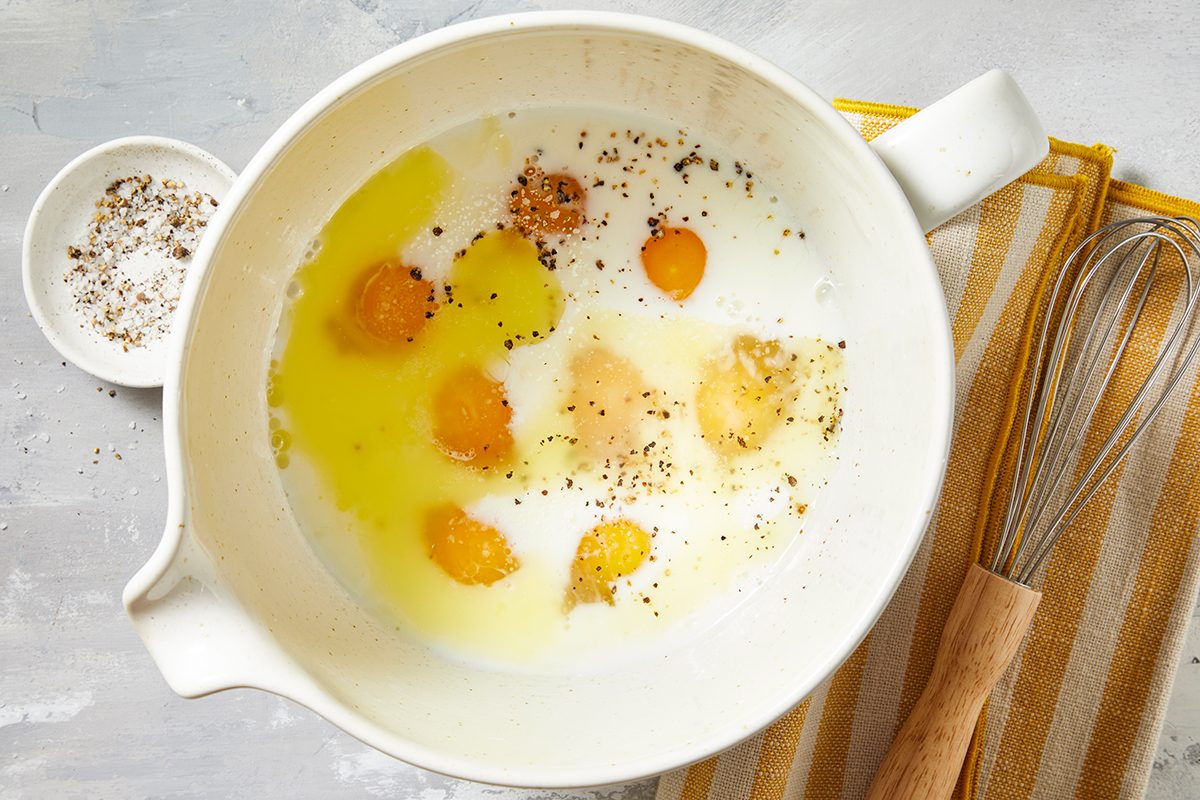 A mixing bowl containing eggs, milk, and pepper, ready for whisking. A wire whisk with a wooden handle lies next to the bowl on a striped yellow and white towel. 