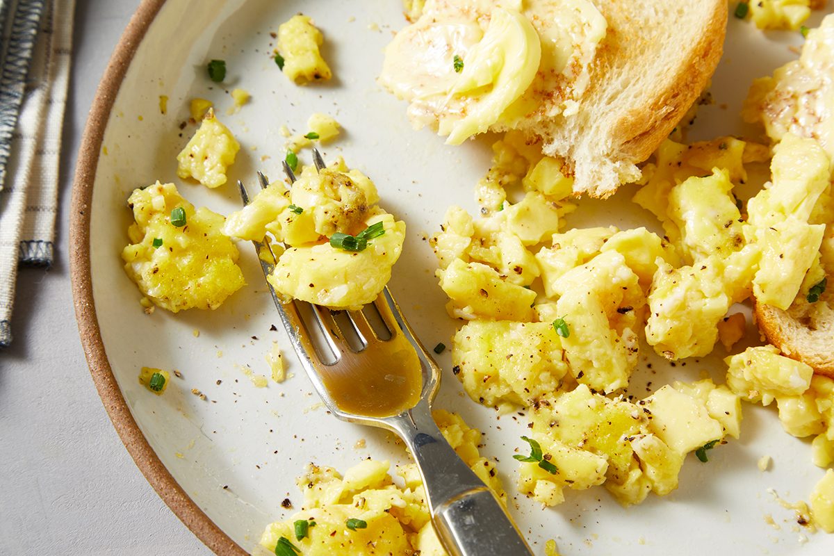 A plate with scrambled eggs seasoned with black pepper and garnished with chopped chives. A fork rests on the plate, and a slice of buttered toast is partially visible.
