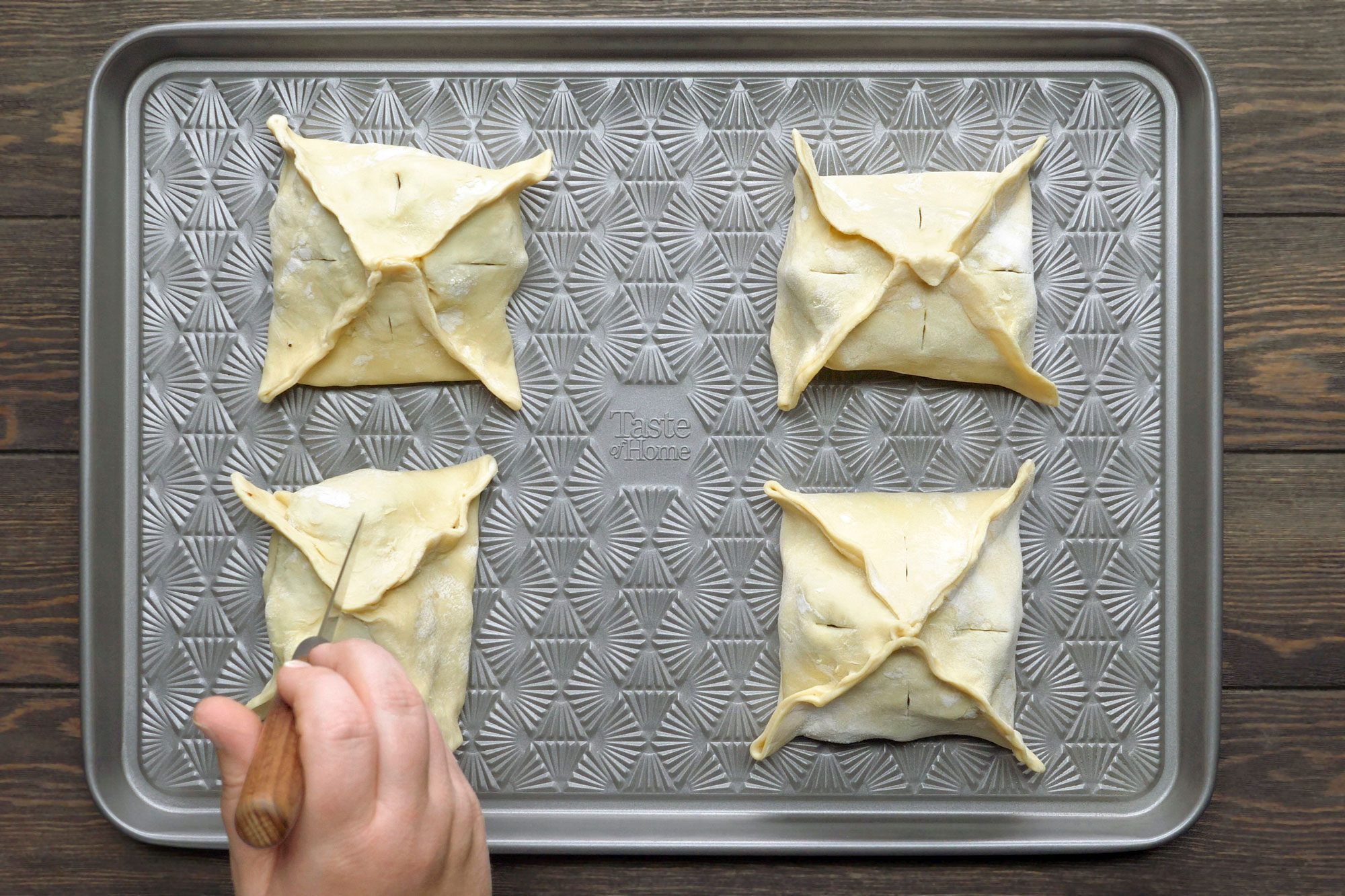overhead shot of a baking sheet with four pastries on it; a hand holds a knife to seal the edge of one pastry; the pastries are in the shape of a square with four points; the baking sheet is gray and has a pattern on it