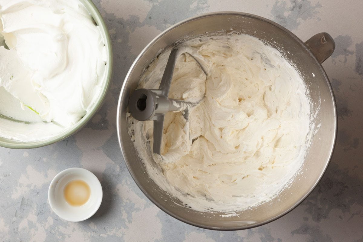 overhead shot of a steel mixing bowl filled frosting or whipped cream, on the left, a light green bowl contains white whipped cream, adjacent to these bowls is a small white dish that holds a liquid, possibly vanilla extract, set against a lightly gray surface