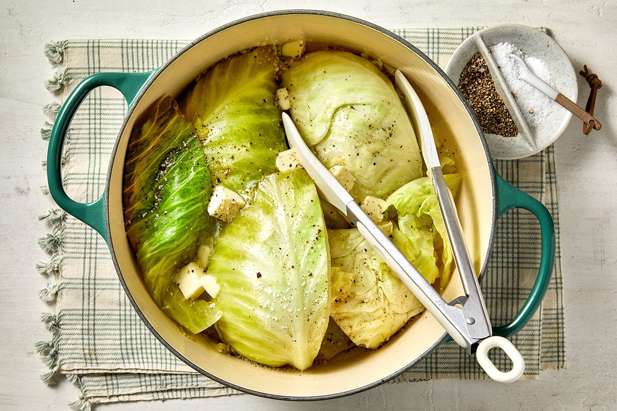A pot filled with cooked cabbage wedges seasoned with pepper and dotted with butter, set on a checkered cloth. Tongs rest in the pot, and a small bowl of pepper and salt is nearby.