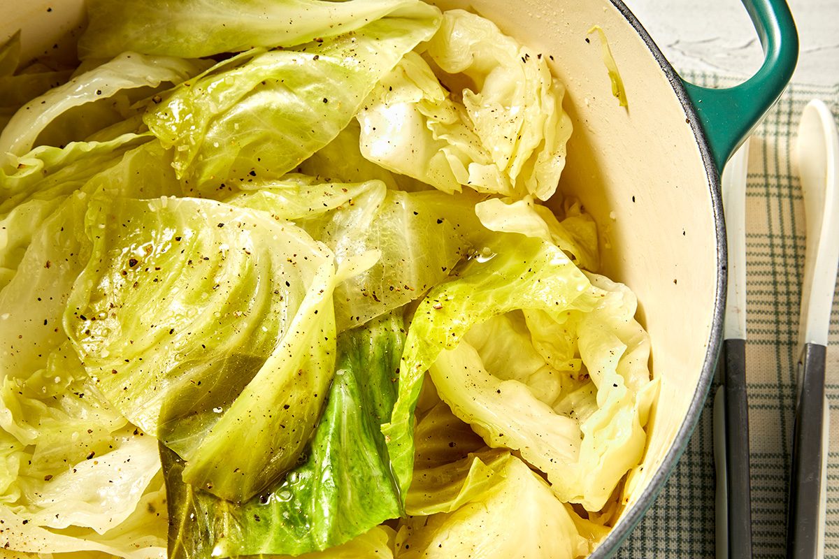 A pot filled with cooked cabbage, seasoned with black pepper, on a green and white plaid tablecloth. Two utensils are laid beside the pot.