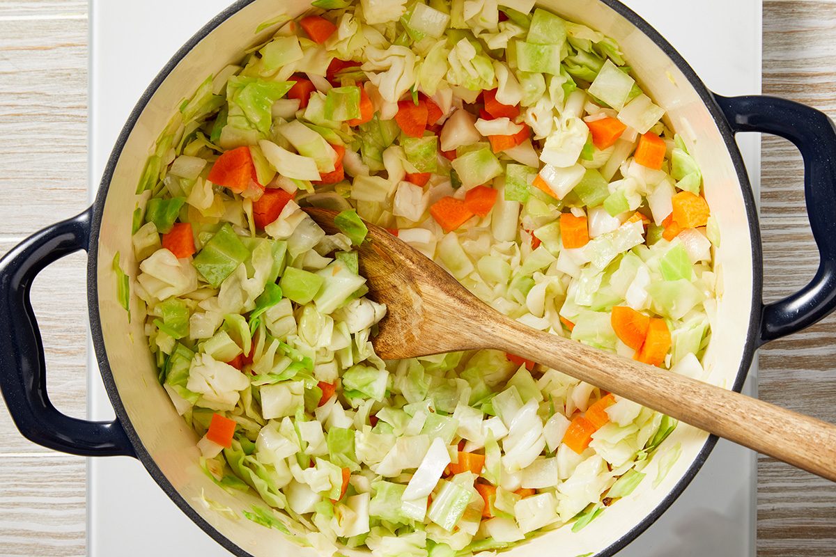 A large pot of chopped cabbage and carrots being stirred with a wooden spoon. The vegetables are brightly colored, with green and orange pieces visible against a light background.