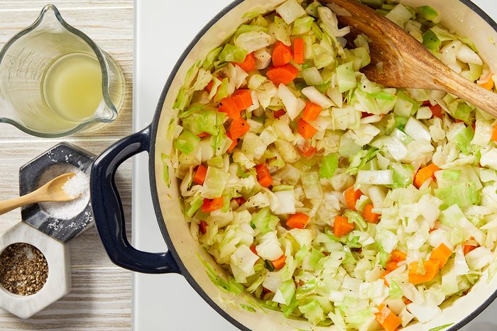 Top view of a pot filled with chopped cabbage, carrots, and onions being stirred with a wooden spoon. To the side, there's a measuring cup with liquid, and small containers with salt and pepper.