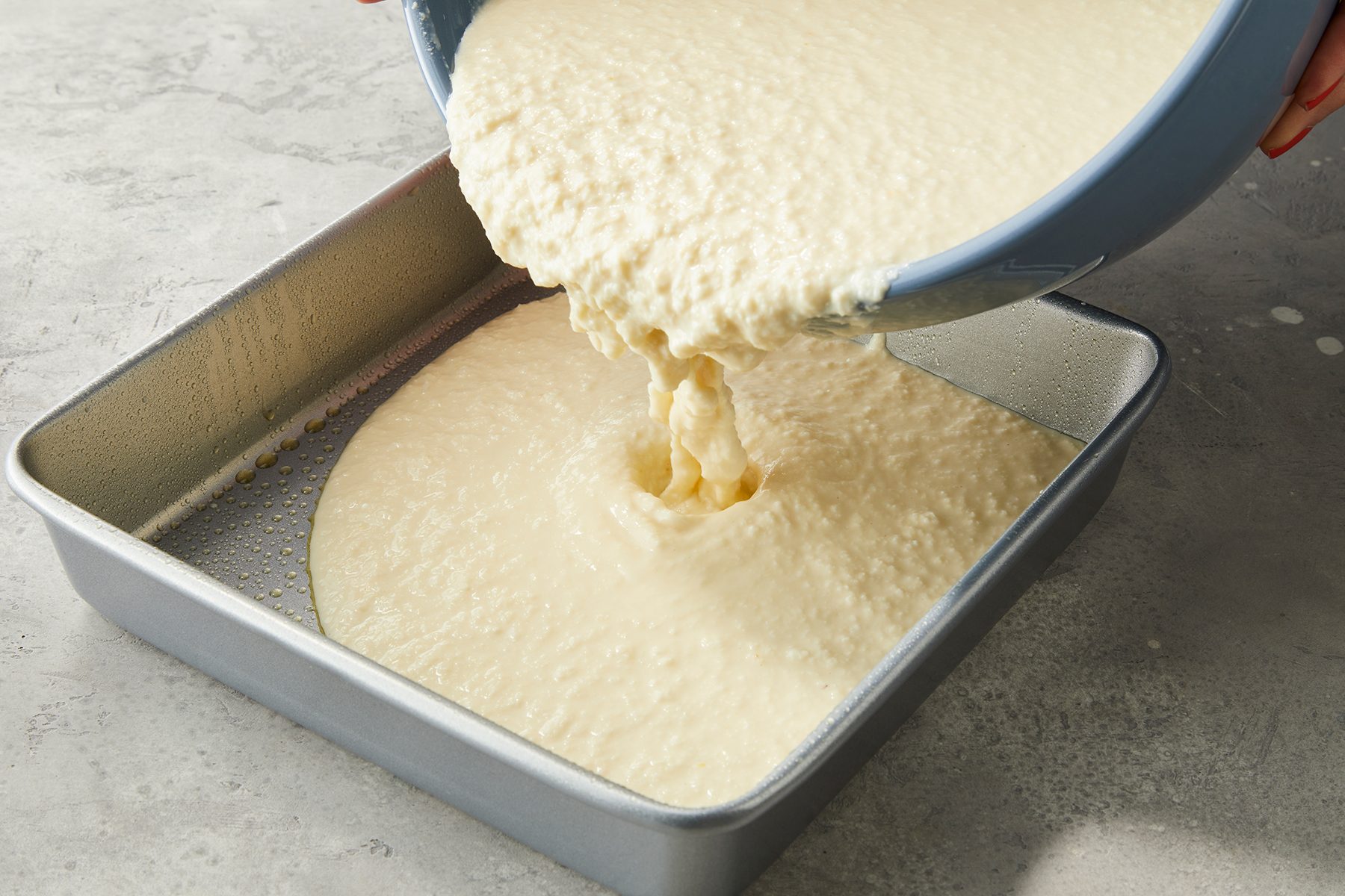 Batter is being poured from a bowl into a greased square baking pan on a gray countertop.