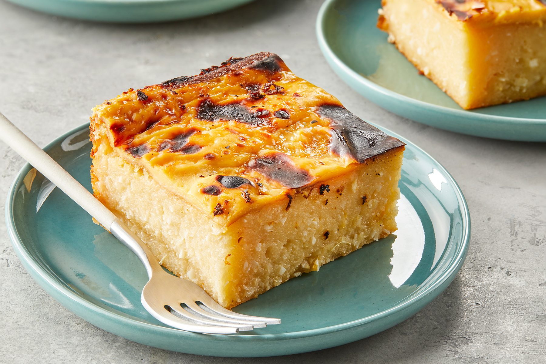 A slice of caramelized cake with a golden brown top sits on a teal ceramic plate. A fork is placed beside the cake on the plate. The background features a similar dish slightly out of focus.