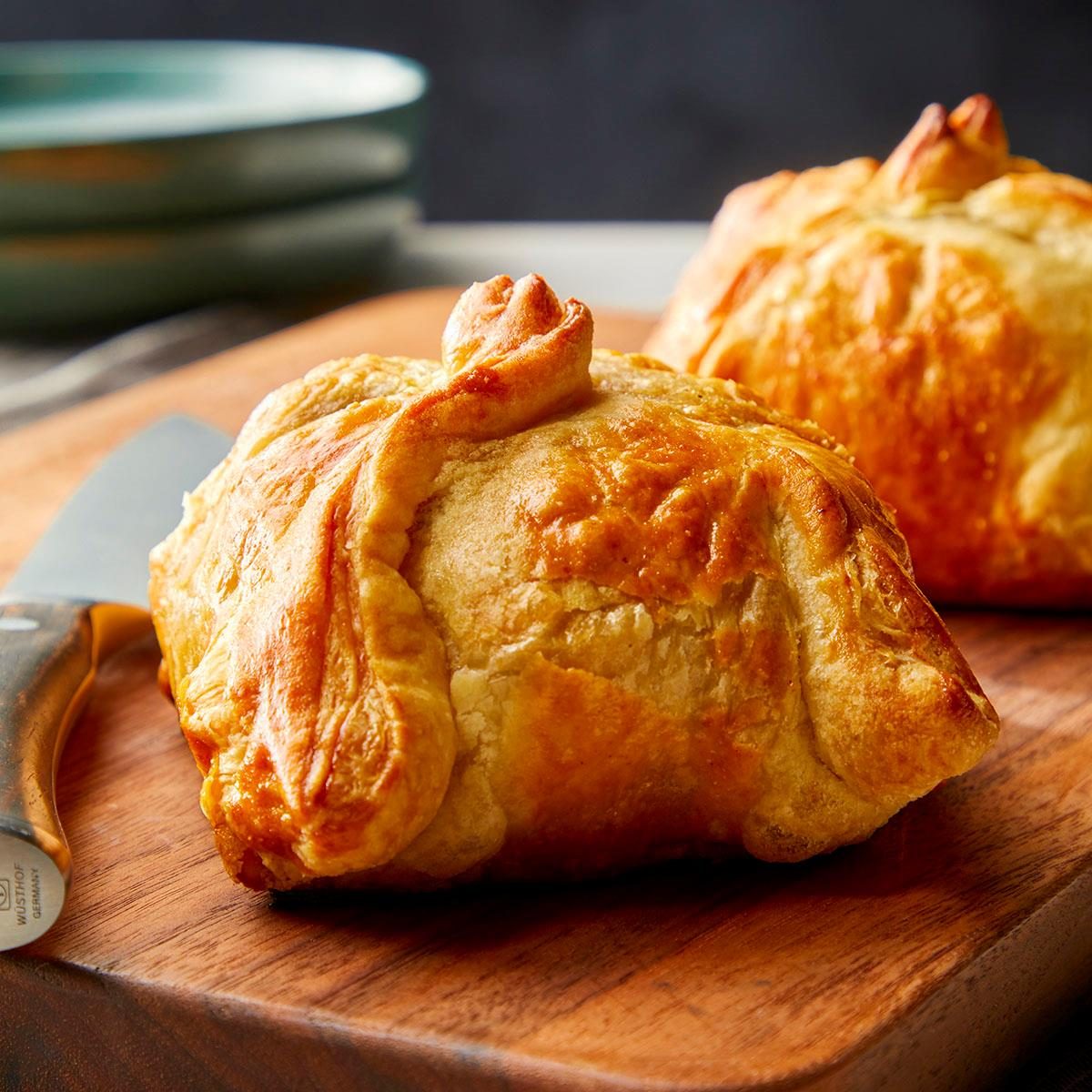 closeup vertical shot of Beef Wellington resting on a wooden cutting board along with a knife; some green plates are visible in background